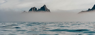 Calm water in the
                foreground, with crags sticking up out of the sea mist.
